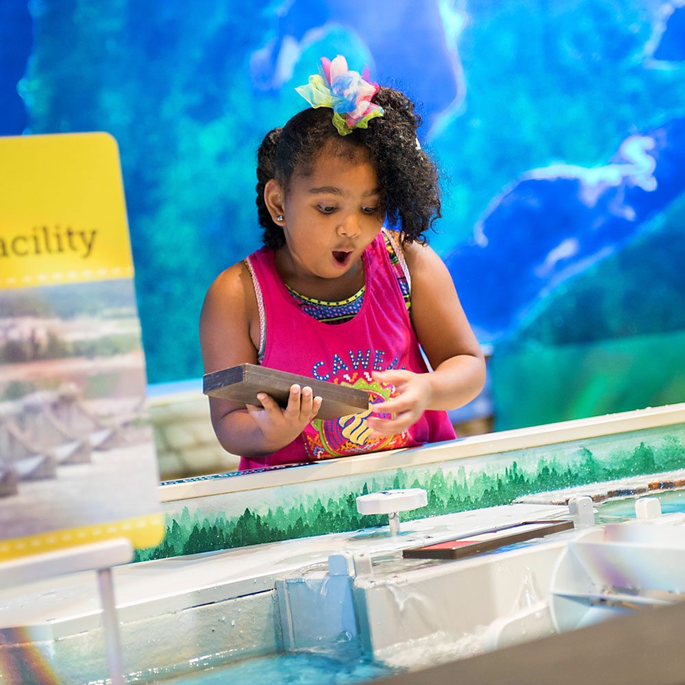 Girl with mouth open in surpise, holding a brown block while standing at the Water Table inside Corteva Agriscience ScienceWorks.